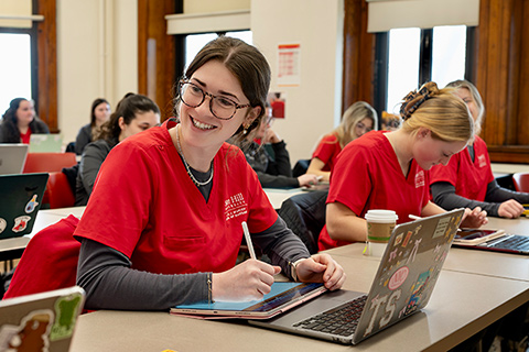 Nursing students wearing red scrubs work on laptops and tablets in a classroom.