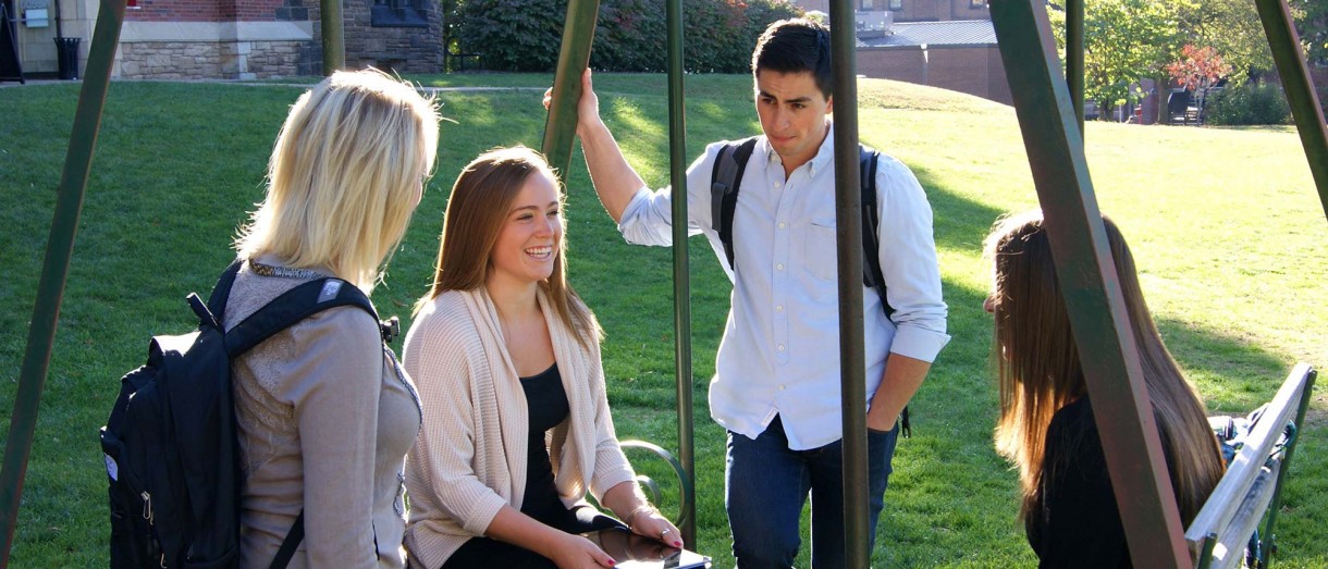 Group of Seton Hill students talking outside on the swings in front of the Sullivan building. 